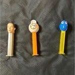 Three vintage and modern Pez dispensers, including a human head character, a clock face, and a blue character on a yellow stem.
