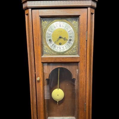 Sale Photo Thumbnail #47: This piece features a fluted wooden case and lower viewing glass showcasing the brass pendulum bob. It was made in West Germany. The key is included. The clock is working at the time of catalog. Measurement: 26.5”H x 12.5”W x 5.5”D. [MI]