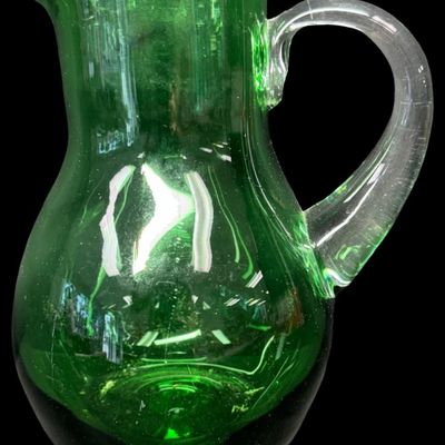 Sale Photo Thumbnail #52: This mid-century hand-blown art glass pitcher is believed to be made by Blenko, or another art glass company of the era. It is emerald green in color, with a clear glass applied handle. There is a rough pontil on the base. There is minor age related wear,