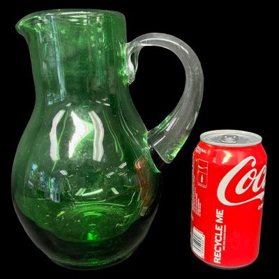 Sale Photo Thumbnail #53: This mid-century hand-blown art glass pitcher is believed to be made by Blenko, or another art glass company of the era. It is emerald green in color, with a clear glass applied handle. There is a rough pontil on the base. There is minor age related wear,