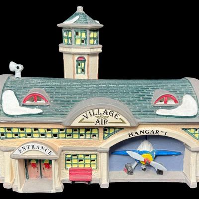 Sale Photo Thumbnail #641: This retired piece features a hand painted ceramic lighted building styled as an airfield hangar, attached control tower, and an integrated single engine prop plane inside. There are no obvious chips or cracks. Measurement: 11” x 8.25”. [AG]