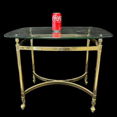 Sale Photo Thumbnail #646: This features a polished brass-finished structure accented by decorative finials at the upper corners, with stylized feet, and a glass top. It is possibly made by Le Barge or Baker. There is no damage to note. Measurement: 27.25”L x 22.25”W x 22”H. 