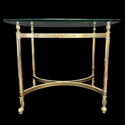 Sale Photo Thumbnail #644: This features a polished brass-finished structure accented by decorative finials at the upper corners, with stylized feet, and a glass top. It is possibly made by Le Barge or Baker. There is no damage to note. Measurement: 27.25”L x 22.25”W x 22”H. 