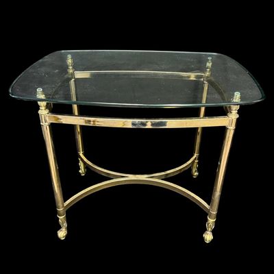Sale Photo Thumbnail #645: This features a polished brass-finished structure accented by decorative finials at the upper corners, with stylized feet, and a glass top. It is possibly made by Le Barge or Baker. There is no damage to note. Measurement: 27.25”L x 22.25”W x 22”H. 