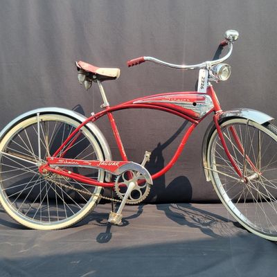 Sale Photo Thumbnail #111: Red Schwinn Jaguar with whitewall tires, full fenders, rear carrier, tank-style frame insert, red Mesinger-style saddle, bell, front light equipment and Jaguar-marked chainguard. The rear wheel sports a Bendix Red Band 2 speed Automatic Hub.  A nicely equ