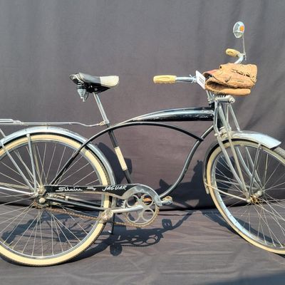 Sale Photo Thumbnail #122: Black Schwinn Jaguar with whitewall tires, full fenders, rear carrier, tank insert, front headlight, rear mud flap and black/white saddle. Attractive survivor with a largely period-correct look and good completeness. Shows scuffs, oxidation and general vi