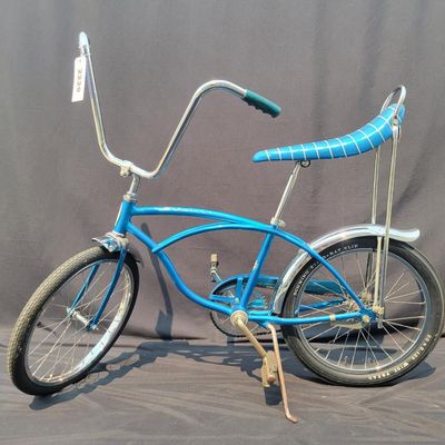 Sale Photo Thumbnail #146: Blue Schwinn Deluxe Sting-Ray single speed coaster brake bicycle with blue banana seat, tall sissy bar, blue grips, high-rise handlebars, fenders and rear reflector. Very complete, original paint and attractive with moderate vintage wear to the saddle, gr