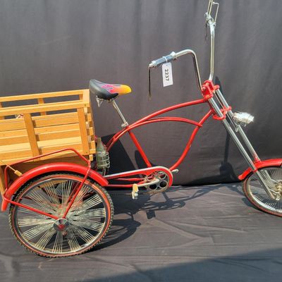 Sale Photo Thumbnail #209: Custom red 3-speed cargo tricycle with long wood-slatted rear cargo bed, tall handlebars, flame-style saddle, headlight, flags and assorted bottle/cargo accessories. Chainguard is marked “Ray Spoiler / 3 Speed.” A fun one-off custom build with strong 