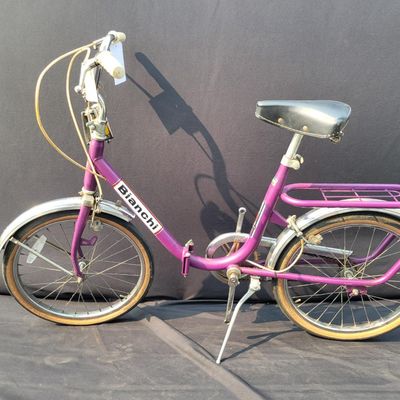Sale Photo Thumbnail #302: Purple/magenta Bianchi folding bicycle with 20-inch wheels, center folding hinge, Bianchi graphics, chrome fenders, rear rack, tan-sidewall tires, high handlebars and twin front light/reflector accessories. Distinctive and substantially complete with grea