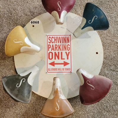 Sale Photo Thumbnail #378: Custom Schwinn-themed display featuring six vintage-style Schwinn “S” saddle tops arranged around a round white base with a red-and-white “SCHWINN PARKING ONLY – ALL OTHERS WILL BE TOWED” center sign. Seat colors include red, teal, yellow, black