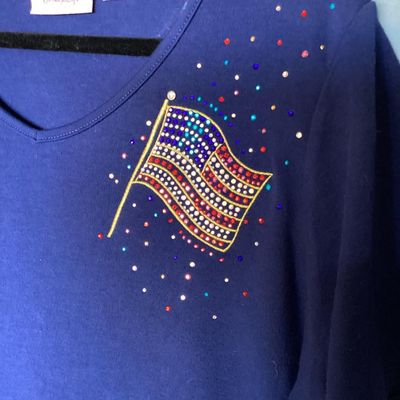 Sale Photo Thumbnail #138: Quacker Factory short sleeve V-neck top in blue, featuring a embellished American flag design on the chest with colorful rhinestones and studs. Tagged size XL. Made in China. Measures approximately 28 inches in length from shoulder to hem. Good pre-owned 
