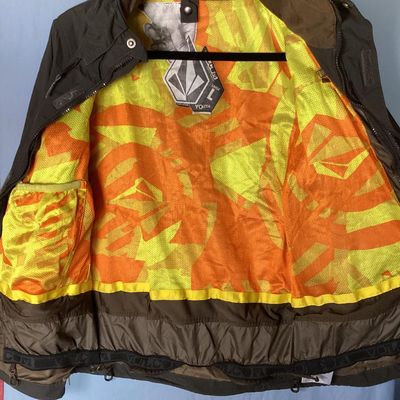 Sale Photo Thumbnail #174: Volcom hooded jacket in dark brown with bright orange and yellow patterned interior lining. Features zip-up and hook-and-loop closures, multiple cargo and zipper pockets, powder skirt, and underarm zip vents. Marked size Youth Large (L). Overall good pre-