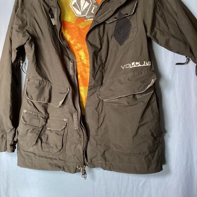 Sale Photo Thumbnail #170: Volcom hooded jacket in dark brown with bright orange and yellow patterned interior lining. Features zip-up and hook-and-loop closures, multiple cargo and zipper pockets, powder skirt, and underarm zip vents. Marked size Youth Large (L). Overall good pre-