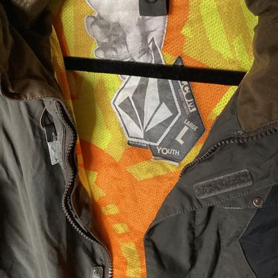 Sale Photo Thumbnail #171: Volcom hooded jacket in dark brown with bright orange and yellow patterned interior lining. Features zip-up and hook-and-loop closures, multiple cargo and zipper pockets, powder skirt, and underarm zip vents. Marked size Youth Large (L). Overall good pre-