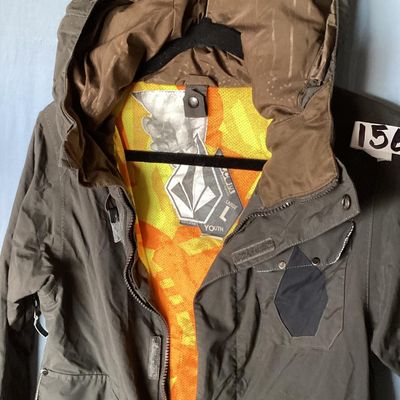 Sale Photo Thumbnail #173: Volcom hooded jacket in dark brown with bright orange and yellow patterned interior lining. Features zip-up and hook-and-loop closures, multiple cargo and zipper pockets, powder skirt, and underarm zip vents. Marked size Youth Large (L). Overall good pre-
