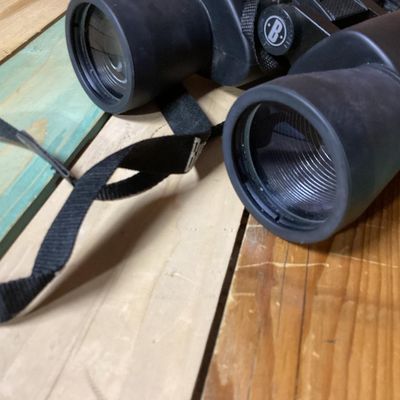 Sale Photo Thumbnail #250: Pair of Bushnell 10x-30x50 zoom binoculars featuring variable magnification controls and a attached black woven neck strap. The casing is finished in a durable black rubberized coating. In used condition with light surface dust and minor wear consistent w