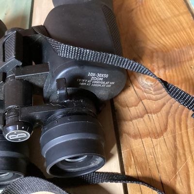 Sale Photo Thumbnail #249: Pair of Bushnell 10x-30x50 zoom binoculars featuring variable magnification controls and a attached black woven neck strap. The casing is finished in a durable black rubberized coating. In used condition with light surface dust and minor wear consistent w