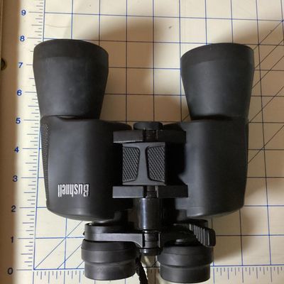 Sale Photo Thumbnail #254: Pair of Bushnell 10x-30x50 zoom binoculars featuring variable magnification controls and a attached black woven neck strap. The casing is finished in a durable black rubberized coating. In used condition with light surface dust and minor wear consistent w