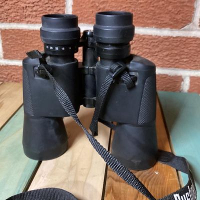 Sale Photo Thumbnail #253: Pair of Bushnell 10x-30x50 zoom binoculars featuring variable magnification controls and a attached black woven neck strap. The casing is finished in a durable black rubberized coating. In used condition with light surface dust and minor wear consistent w