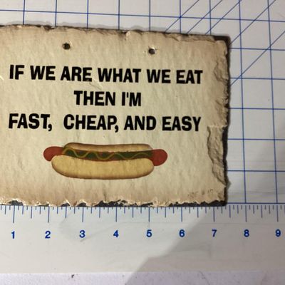 Sale Photo Thumbnail #266: A slate hanging wall plaque featuring the humorous phrase: "IF WE ARE WHAT WE EAT THEN I'M FAST, CHEAP, AND EASY" above a hot dog graphic. Signed/dated CNE '97 on the front lower corner. Features prepunched holes at the top for hanging and a rough deckled