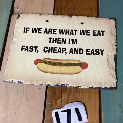 Sale Photo Thumbnail #262: A slate hanging wall plaque featuring the humorous phrase: "IF WE ARE WHAT WE EAT THEN I'M FAST, CHEAP, AND EASY" above a hot dog graphic. Signed/dated CNE '97 on the front lower corner. Features prepunched holes at the top for hanging and a rough deckled