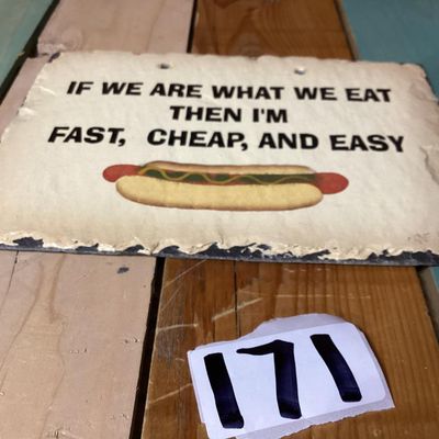 Sale Photo Thumbnail #263: A slate hanging wall plaque featuring the humorous phrase: "IF WE ARE WHAT WE EAT THEN I'M FAST, CHEAP, AND EASY" above a hot dog graphic. Signed/dated CNE '97 on the front lower corner. Features prepunched holes at the top for hanging and a rough deckled