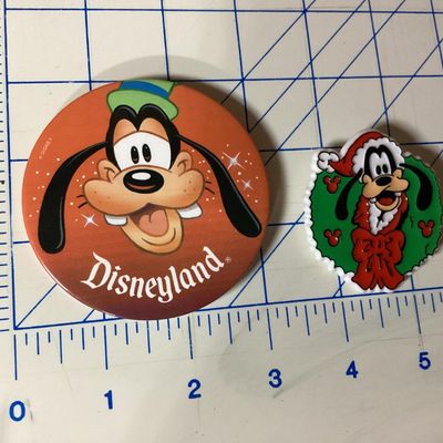 Sale Photo Thumbnail #270: Pair of vintage Goofy collectible pins/brooches featuring:
- A large circular Disneyland button pin featuring Goofy's face, measuring approximately 3 inches in diameter. Has light adhesive residue on the back.
- A plastic/molded Christmas wreath brooch fe