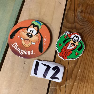Sale Photo Thumbnail #267: Pair of vintage Goofy collectible pins/brooches featuring:
- A large circular Disneyland button pin featuring Goofy's face, measuring approximately 3 inches in diameter. Has light adhesive residue on the back.
- A plastic/molded Christmas wreath brooch fe