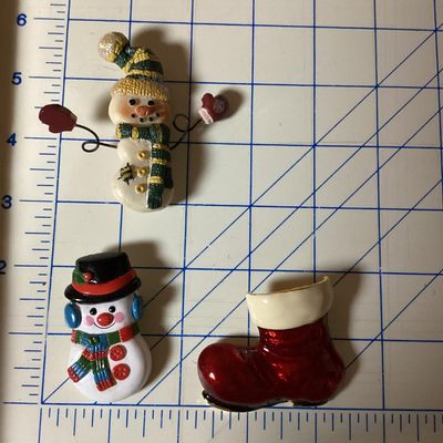Sale Photo Thumbnail #274: A festive collection of three Christmas-themed brooches and pins. Includes a red enameled Santa boot/stocking brooch, a painted plastic snowman wearing a black top hat, and a textured resin/painted snowman pin with wire-articulated mitten arms. Each pin m