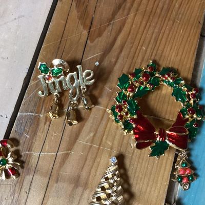 Sale Photo Thumbnail #278: Collection of festive Christmas-themed fashion jewelry, including:
- Enamel wreath brooch with red bow accent and dangling Christmas tree
- Gold-tone 'Jingle' pin with dangling bell charms
- Gold-tone woven Christmas tree brooch with rhinestone top
- Two 
