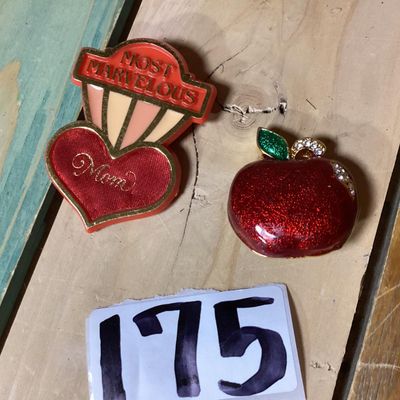 Sale Photo Thumbnail #287: Pair of vintage brooches including:
- 1983 Hallmark Cards, Inc. plastic/enamel heart pin that reads "Most Marvelous Mom". Measures approximately 2.25 inches tall.
- Red glitter enamel and rhinestone gold-tone apple brooch. Measures approximately 1.25 inch