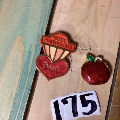 Sale Photo Thumbnail #286: Pair of vintage brooches including:
- 1983 Hallmark Cards, Inc. plastic/enamel heart pin that reads "Most Marvelous Mom". Measures approximately 2.25 inches tall.
- Red glitter enamel and rhinestone gold-tone apple brooch. Measures approximately 1.25 inch
