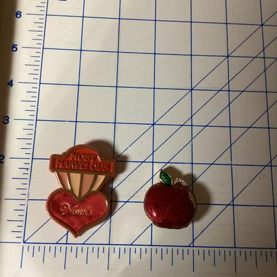 Sale Photo Thumbnail #285: Pair of vintage brooches including:
- 1983 Hallmark Cards, Inc. plastic/enamel heart pin that reads "Most Marvelous Mom". Measures approximately 2.25 inches tall.
- Red glitter enamel and rhinestone gold-tone apple brooch. Measures approximately 1.25 inch