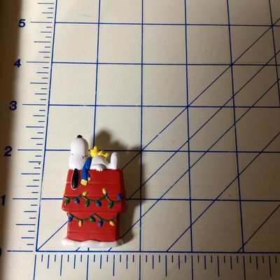 Sale Photo Thumbnail #293: Vintage Hallmark Cards Peanuts collectible lapel pin featuring Snoopy and Woodstock sleeping atop a red doghouse decorated with Christmas lights. Marked on back '© UFS HMK. CDS.' Pin clasp on back intact. Measures approximately 2 inches tall. In good pre