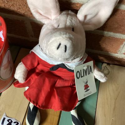 Sale Photo Thumbnail #327: Collection of vintage Olivia the Pig items featuring a GUND plush doll wearing a red dress with original tags attached and a matching Schylling red tin pail with handle and original tag attached (dated 2003). Both items are in good overall condition with 