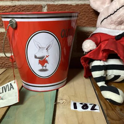 Sale Photo Thumbnail #326: Collection of vintage Olivia the Pig items featuring a GUND plush doll wearing a red dress with original tags attached and a matching Schylling red tin pail with handle and original tag attached (dated 2003). Both items are in good overall condition with 