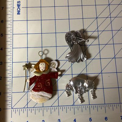 Sale Photo Thumbnail #351: Lot of 3 decorative brooches featuring an angel with a trumpet, a painted angel holding a wand/star, and a silver-tone pig figure. Each piece measures approximately 2 to 4 inches long. Pre-owned condition with light surface wear visible.