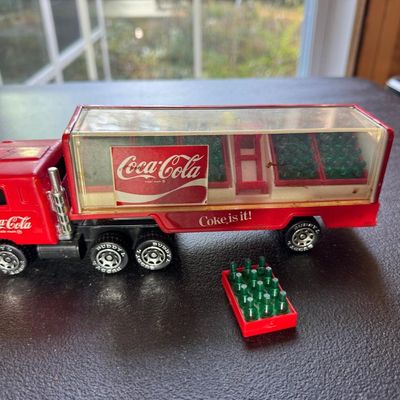 Sale Photo Thumbnail #120: Vintage Buddy L Coca-Cola delivery semi truck, red cab and trailer with clear display sides, "Coca-Cola" and "Coke is it!" graphics, Buddy L-marked wheels, and miniature red bottle cases with green bottles (one loose case shown).
Condition: Played-with; s