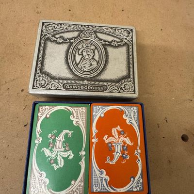 Sale Photo Thumbnail #133: Vintage Gainsborough double-deck bridge set: green and orange decks with ornate scroll and cornucopia backs, in the original embossed silver-tone presentation box with cameo portrait lid.
Condition: Light box wear. Decks not counted for completeness.

Sol