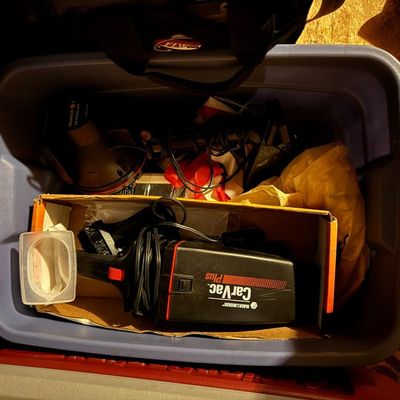 Sale Photo Thumbnail #181: Storage bin of car and garage accessories, including a Black & Decker CarVac Plus 12V car vacuum with attachments and original box, a Sunspot "The Power Light" handheld spotlight, cords, a tire gauge, first-aid items, and assorted accessories. Storage tot