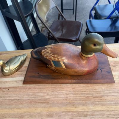 Sale Photo Thumbnail #2: Lot of two duck collectibles featuring a hand-painted carved wood mallard decoy mounted on a wooden display base alongside a small solid brass duck figurine. Both pieces show light surface wear consistent with age and display, including slight scuffs to t