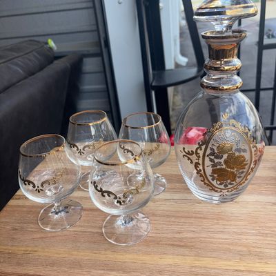 Sale Photo Thumbnail #14: A vintage clear glass decanter set featuring ornate gold-tone scrollwork and floral medallion designs. Includes one decanter with a matching stopper and four small snifter-style glasses with gold rims and decorative scroll accents. Appears in good pre-own