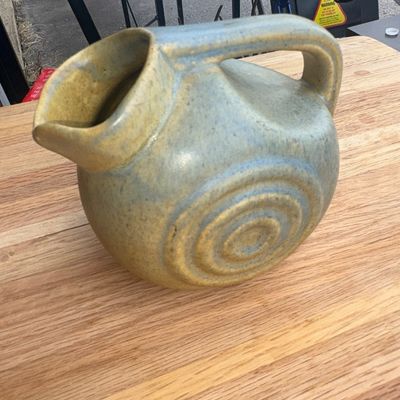 Sale Photo Thumbnail #19: A vintage Art Deco era ceramic pitcher by Nilak Pottery featuring a rounded form with relief concentric circles on the sides and a integrated handle. Finished in a matte yellow/mustard and grey glaze blend. Marked 'NILAK' on the bottom along with a mold n