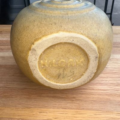 Sale Photo Thumbnail #20: A vintage Art Deco era ceramic pitcher by Nilak Pottery featuring a rounded form with relief concentric circles on the sides and a integrated handle. Finished in a matte yellow/mustard and grey glaze blend. Marked 'NILAK' on the bottom along with a mold n