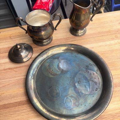 Sale Photo Thumbnail #21: A 4-piece vintage silver plate service set including a creamer pitcher, double-handled sugar bowl with lid, and a round serving tray. Items show heavy tarnishing, patina, and surface wear consistent with age and use. Ideal for polishing or decorative disp