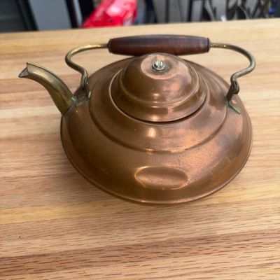 Sale Photo Thumbnail #22: A vintage metal tea kettle featuring a copper body, brass spout, and a overhead handle with a wooden grip. Shows light surface tarnish, patina, and minor surface wear consistent with age and use large dent