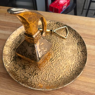 Sale Photo Thumbnail #23: A vintage gold weeping gold glaze decorative pitcher and matching round tray, accompanied by a small brass bottle opener. The tray features a textured mottling pattern typical of mid-century weeping gold ceramic artware. Shows light age-appropriate wear a