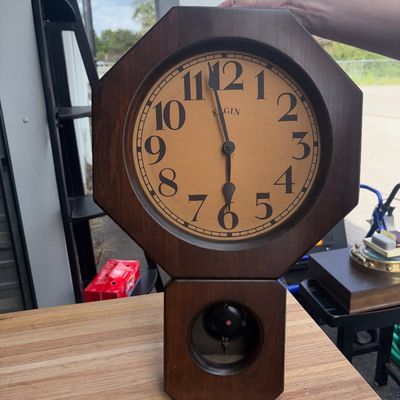Sale Photo Thumbnail #34: Vintage wooden wall clock by Elgin featuring an octagonal head and lower pendulum window. Shows light surface wear and minor scuffs to the wood finish consistent with age and use. Clock mechanism and functionality are unverified.