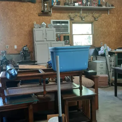 Workshop and garage area filled with various items including a floor jack, wooden coffee tables, a blue storage tote, cabinets, and...