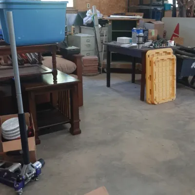 Workshop space filled with various items including a hydraulic floor jack, wooden end tables, plastic storage totes, and automotive parts...